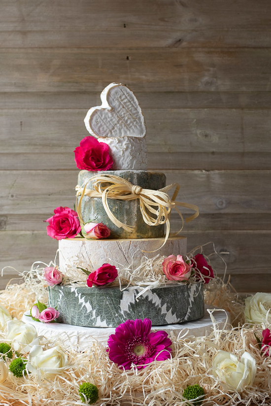 5 tiers of various types of cheese with decorative pink, red and green flowers and a light ivory coloured raffia.