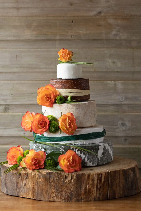 The Ava Wedding Cheese cake has 5 tiers with beautiful orange decorative roses surrounding the cake. two of the layers is wrapped in forest green and ivory raffia.